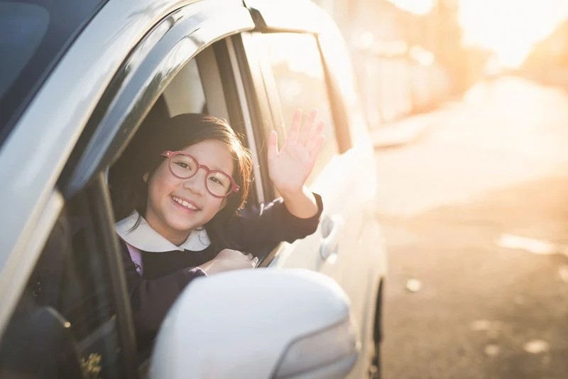 A smiling female Asian child, wearing MiyoSmart lenses, waving in the passenger seat of a car
